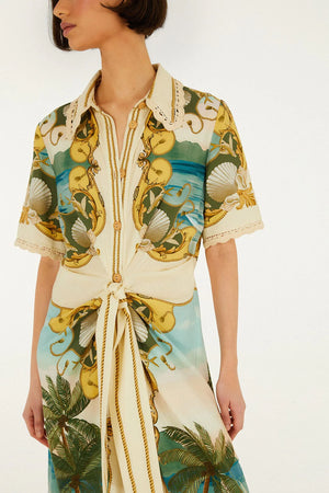 A woman wears the Royal Sea Multicolor Midi Dress by Farm Rio, showcasing its short sleeves and tropical seashell and palm tree print with gold accents. She stands against a plain background, emphasizing the dress’s vibrant nature-inspired design.
