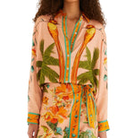 A woman wears a peach Birds Canto Dos Passaros Shirt by FARM Rio with palm tree prints, paired with a matching floral skirt featuring a tie waist.