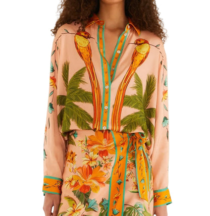 A woman wears a peach Birds Canto Dos Passaros Shirt by FARM Rio with palm tree prints, paired with a matching floral skirt featuring a tie waist.