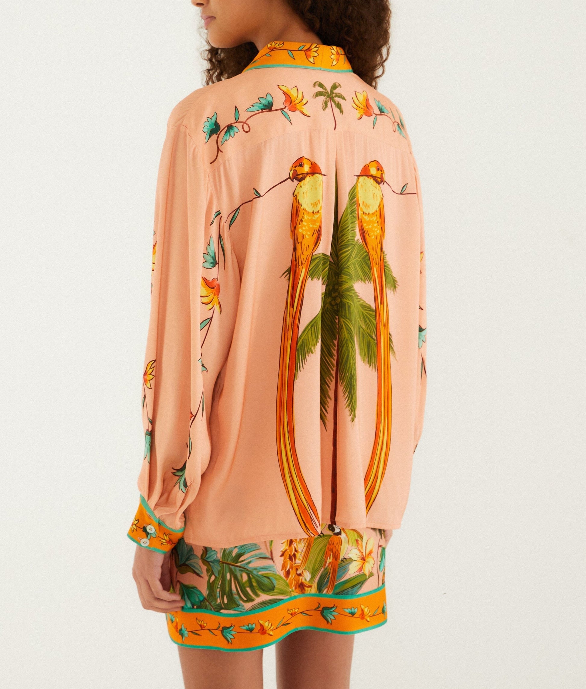 A person wearing a Farm Rio Birds Canto Dos Passardos Shirt, peach-colored with two long-tailed birds and green leaves printed on the back, paired with a matching floral skirt.