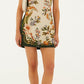 A woman wears a sleeveless Nature Sand Mini Dress by Farm Rio with a floral pattern, green side panels, styled with a white shoulder bag and beige flats.