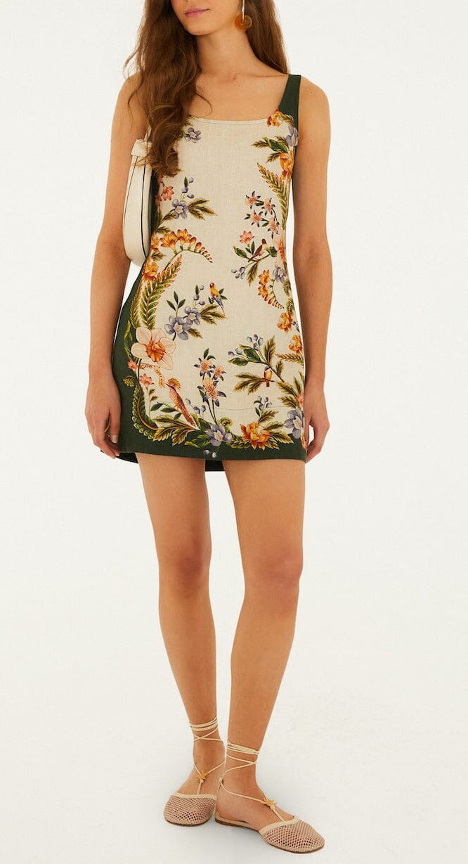 A woman wears a sleeveless Nature Sand Mini Dress by Farm Rio with a floral pattern, green side panels, styled with a white shoulder bag and beige flats.
