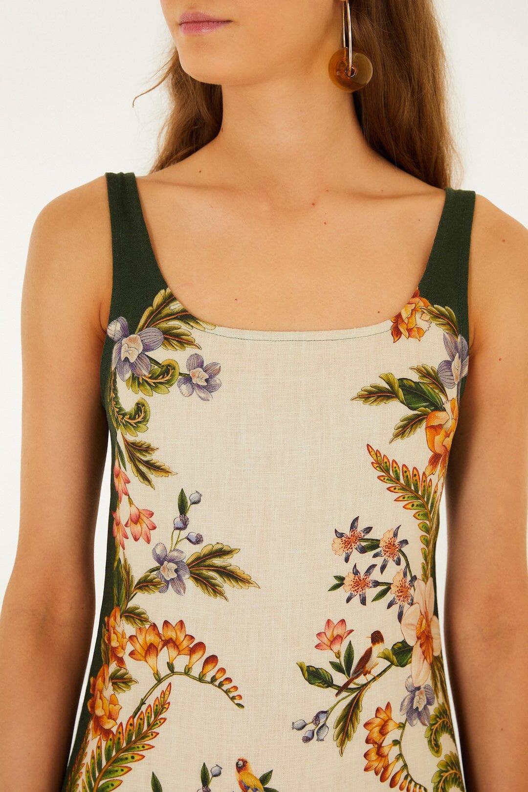A woman with long brown hair and circular earrings wears the Farm Rio Nature Sand Mini Dress, featuring a floral print and green straps.