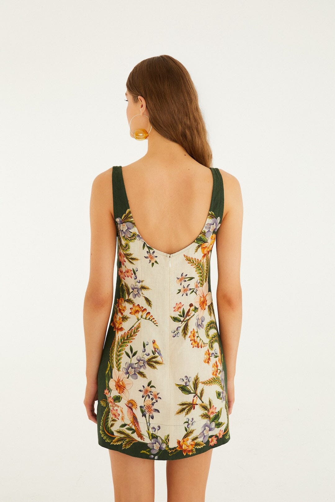 A woman with long brown hair wears the Farm Rio Nature Sand Mini Dress, a sleeveless floral mini with a square neck and scoop back, as she stands against a plain white background.