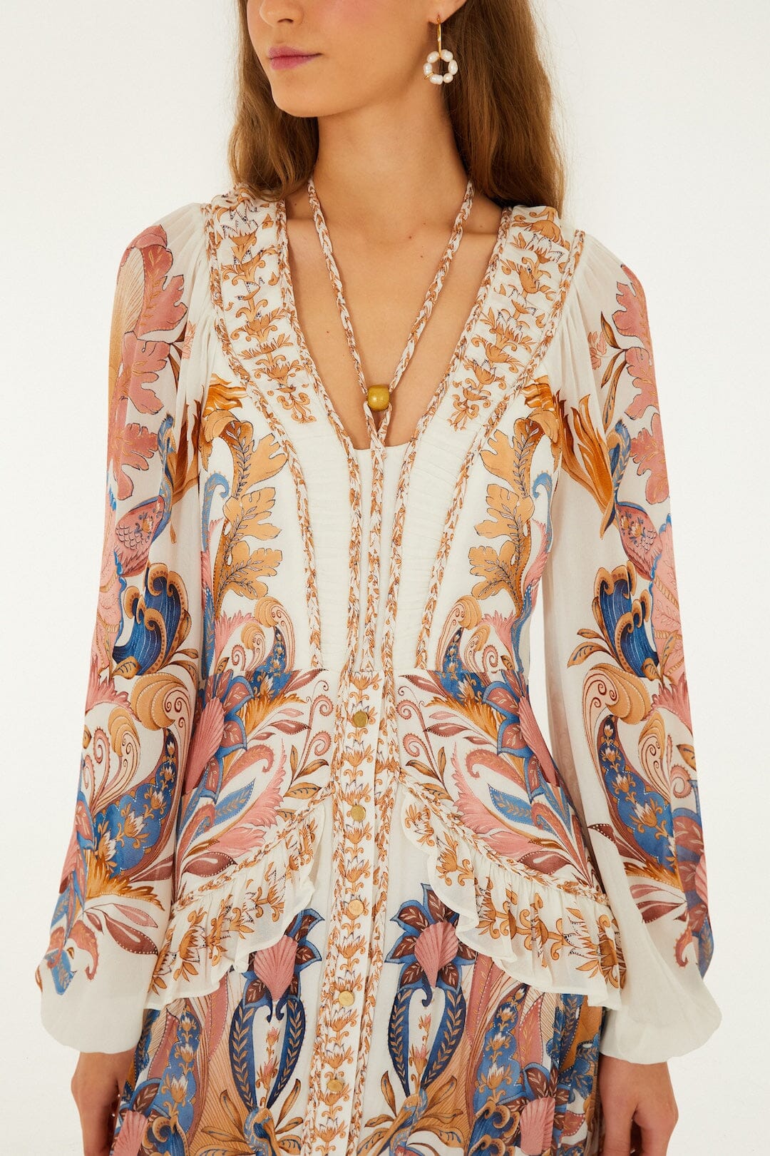A woman wearing the Farm Rio Sea Garden Off White Braid Details Long Dress, featuring floral and paisley designs in pink, blue, and gold, accessorized with a necklace and drop earrings.