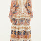 A woman faces away, wearing the Farm Rio Sea Garden Off White Braid Details Long Dress with long sleeves and ruffled layers, styled with flat sandals.