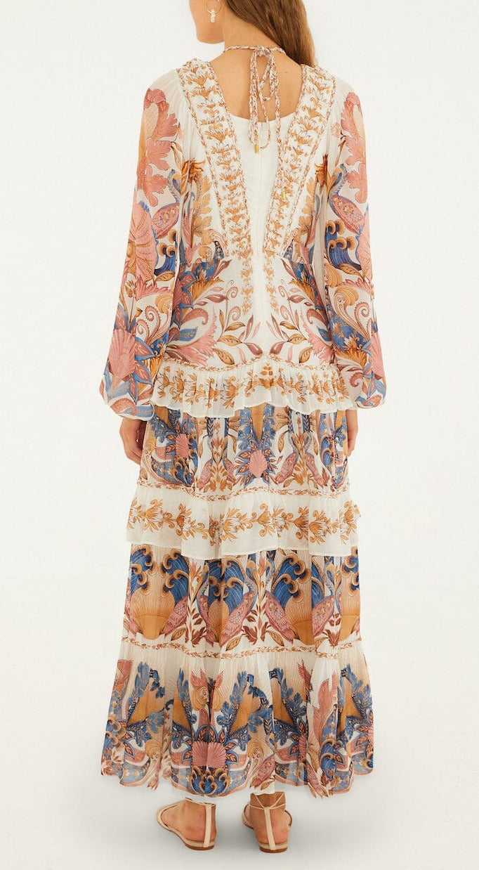 A woman faces away, wearing the Farm Rio Sea Garden Off White Braid Details Long Dress with long sleeves and ruffled layers, styled with flat sandals.