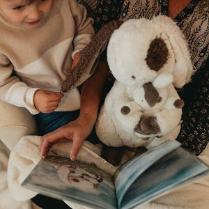A child and grandparent share a sweet moment with Bunnies By the Bay’s stuffed dog toy while reading "What Will My Grandchild Call Me?," a storybook featuring images of a similar dog—a special keepsake time together.