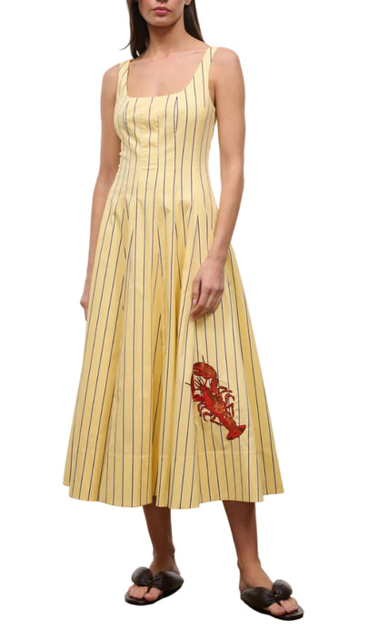 A woman wears the Staud Wells Monday Stripe Lobster Dress—a sleeveless yellow cotton poplin dress with thin vertical blue stripes, fitted waist, and red lobster graphic—paired with black slides.