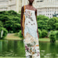 A woman wears a Farm Rio Toile De Rio Off White Maxi Dress, a sleeveless cream garment featuring a landscape print with mountains, birds, and flowers.