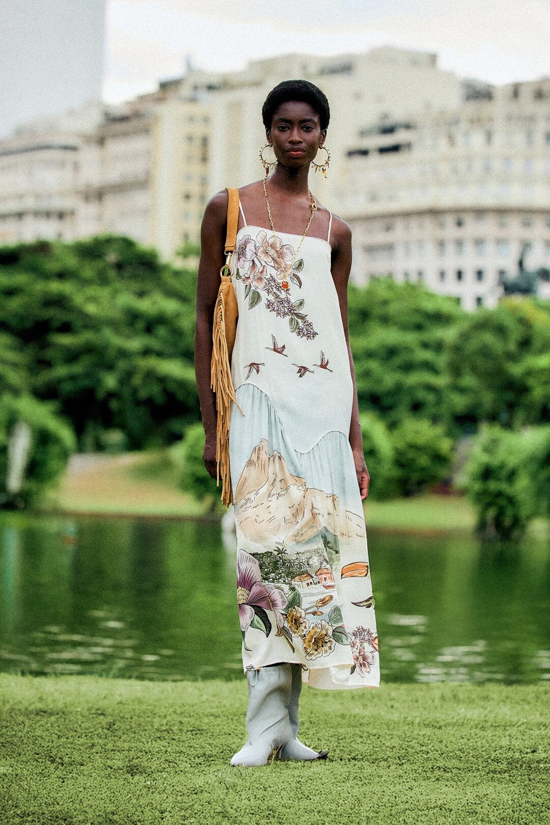 A woman wears a Farm Rio Toile De Rio Off White Maxi Dress, a sleeveless cream garment featuring a landscape print with mountains, birds, and flowers.
