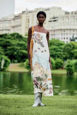 A woman wears a Farm Rio Toile De Rio Off White Maxi Dress, a sleeveless cream garment featuring a landscape print with mountains, birds, and flowers.