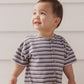 A toddler wearing the Rylee & Cru Baby Boys’ Hayes Jumpsuit with blue and white stripes and coconut buttons stands in front of a white wooden wall, looking upward and smiling.