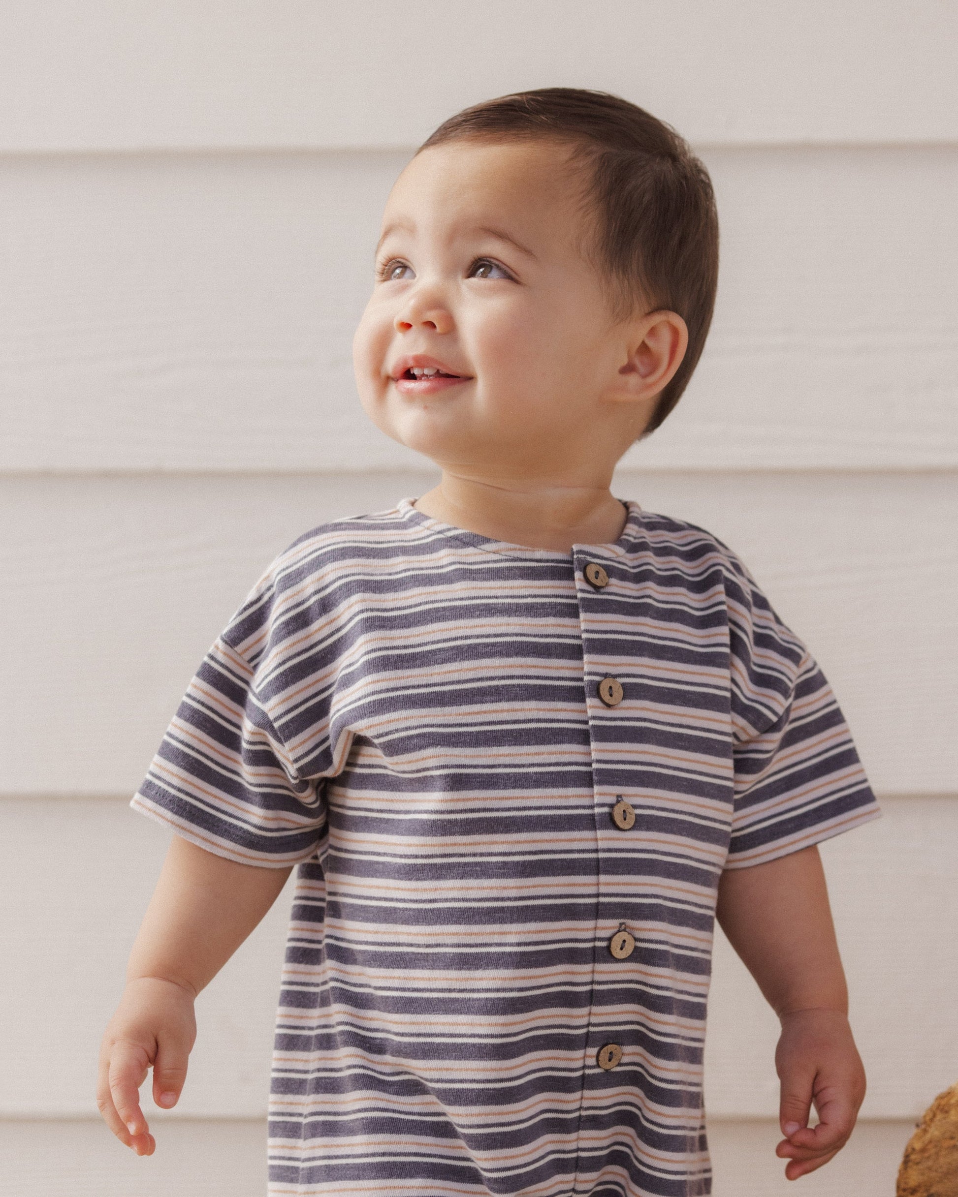 A toddler wearing the Rylee & Cru Baby Boys’ Hayes Jumpsuit with blue and white stripes and coconut buttons stands in front of a white wooden wall, looking upward and smiling.