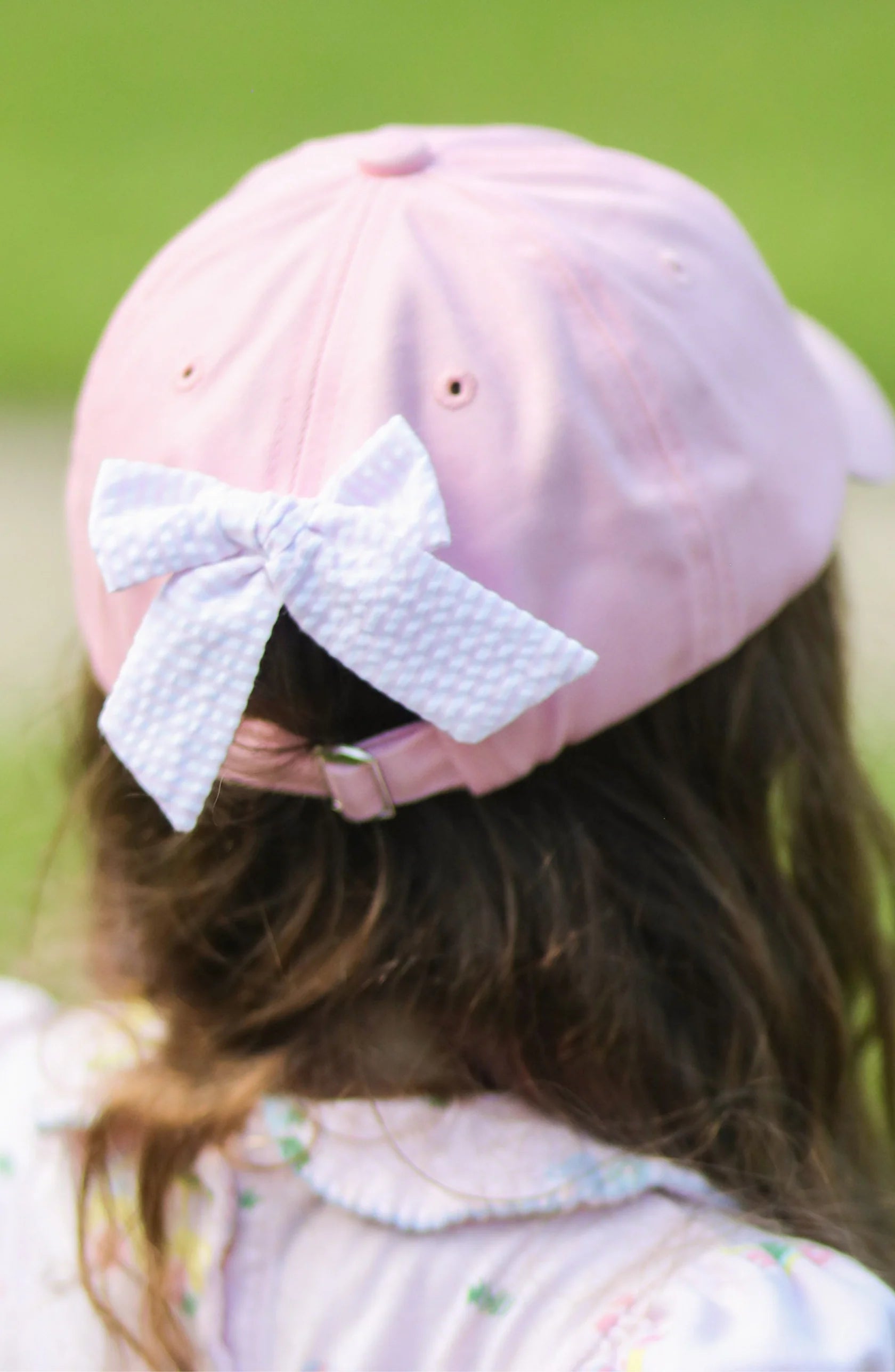 A child is seen from behind wearing the Bits & Bows Rainbow Golf Cart Bow Baseball Hat—a pastel pink girls’ accessory with a patterned bow at the back, perfect for sun-shielding sportswear.