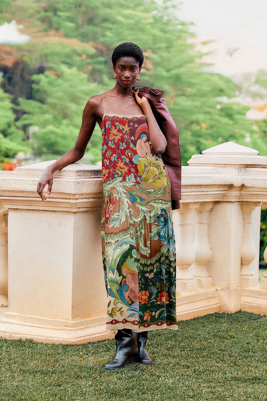 A person wearing a Lunar Boho Maxi Dress, boots, and a jacket draped over one shoulder while leaning against a stone railing.