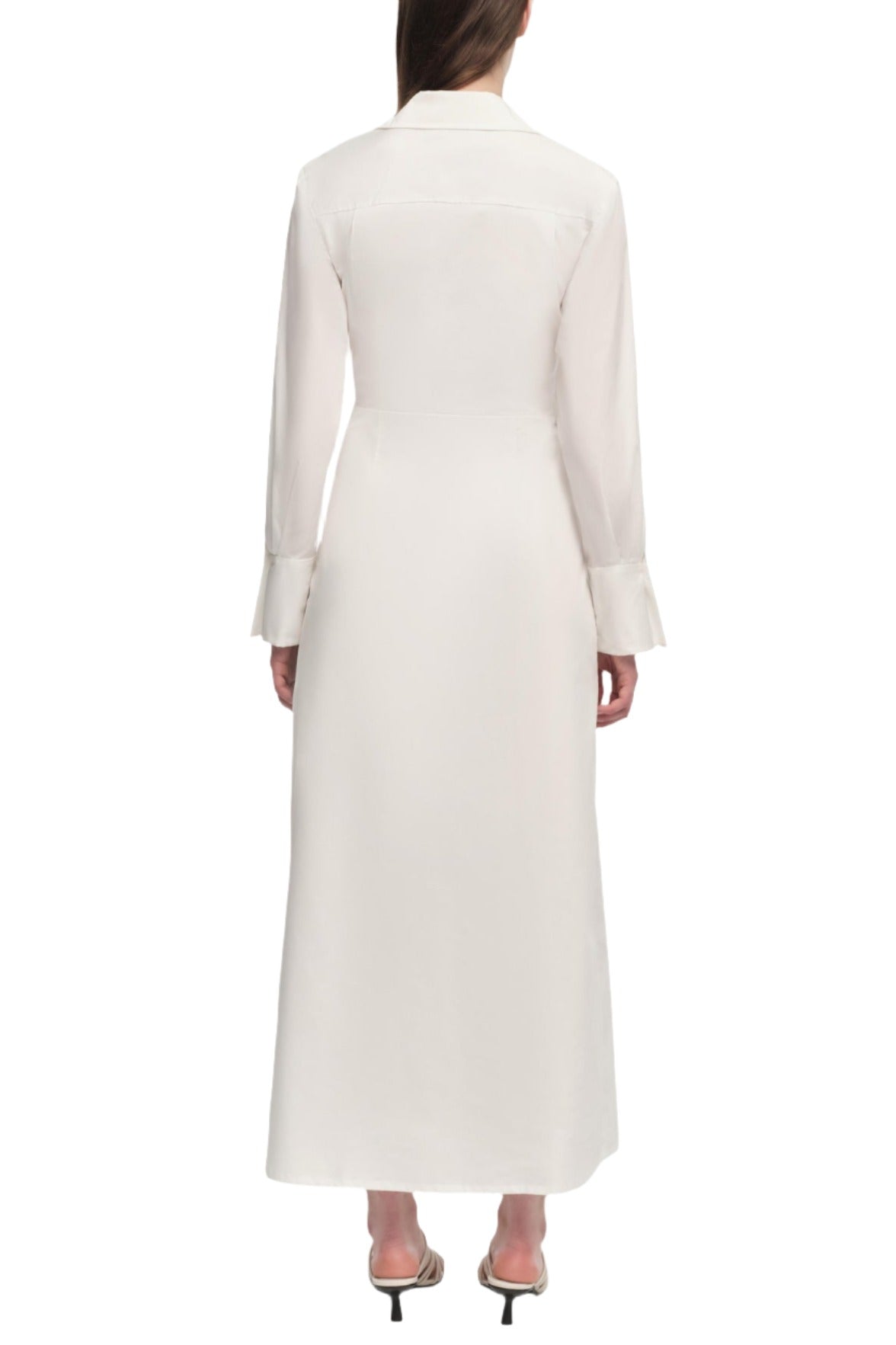 Back view of a person wearing the Simkhai Briar Dress, a long white wrap-style front dress with long sleeves and a collar. The hemline reaches the ankles, and the person is wearing white high-heeled shoes.