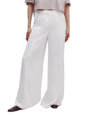 Someone stands against a plain white background wearing Dee's Trouser by TWP, a relaxed high-rise wide leg pant in white, paired with a light-colored shirt and dark pointed shoes.
