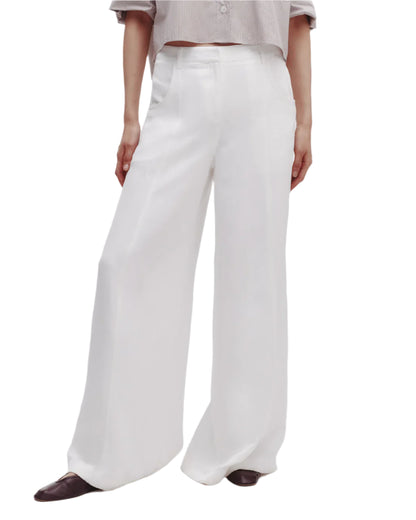 Someone stands against a plain white background wearing Dee's Trouser by TWP, a relaxed high-rise wide leg pant in white, paired with a light-colored shirt and dark pointed shoes.