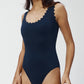 A woman models the Karla Colletto Ines Round Neck One Piece, a navy swimsuit featuring rick rack edging on the neckline and armholes, set against a plain background.