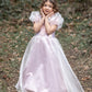A young girl wears the Great Pretenders Platinum Princess Gown, featuring light pink puffy sleeves, as she stands outdoors on a leaf-covered path with her hands near her face and greenery in the background.