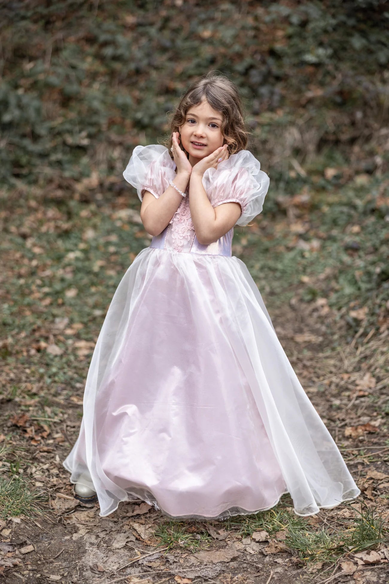 A young girl wears the Great Pretenders Platinum Princess Gown, featuring light pink puffy sleeves, as she stands outdoors on a leaf-covered path with her hands near her face and greenery in the background.