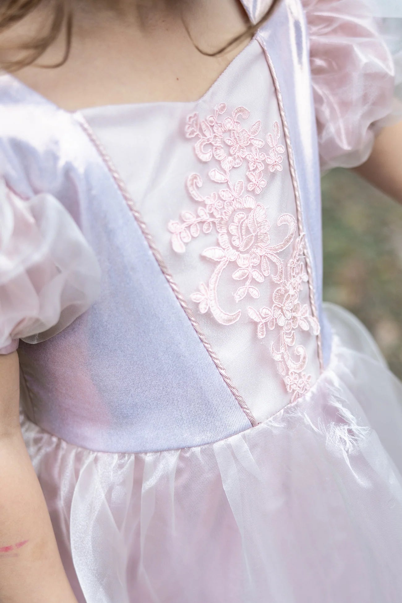 A child wearing the Great Pretenders Platinum Princess Gown, a light pink kids’ dress featuring floral embroidery and sheer puff sleeves, shown from the shoulders to the waist.