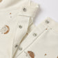 Close-up of the Rylee & Cru Baby Raglan Jumpsuit, a cream-colored French terry romper adorned with metal snap buttons and a delightful pattern of small hedgehogs and mushrooms.