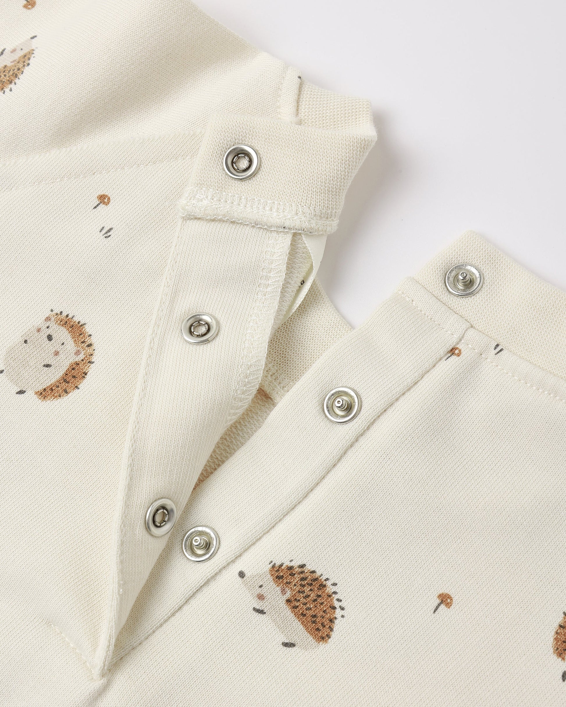 Close-up of the Rylee & Cru Baby Raglan Jumpsuit, a cream-colored French terry romper adorned with metal snap buttons and a delightful pattern of small hedgehogs and mushrooms.