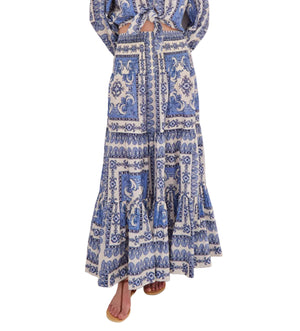 A person in the Cara Cara Emilia Skirt—a blue and white placed print smocked maxi skirt—and matching top, barefoot in sandals, stands against a white background.