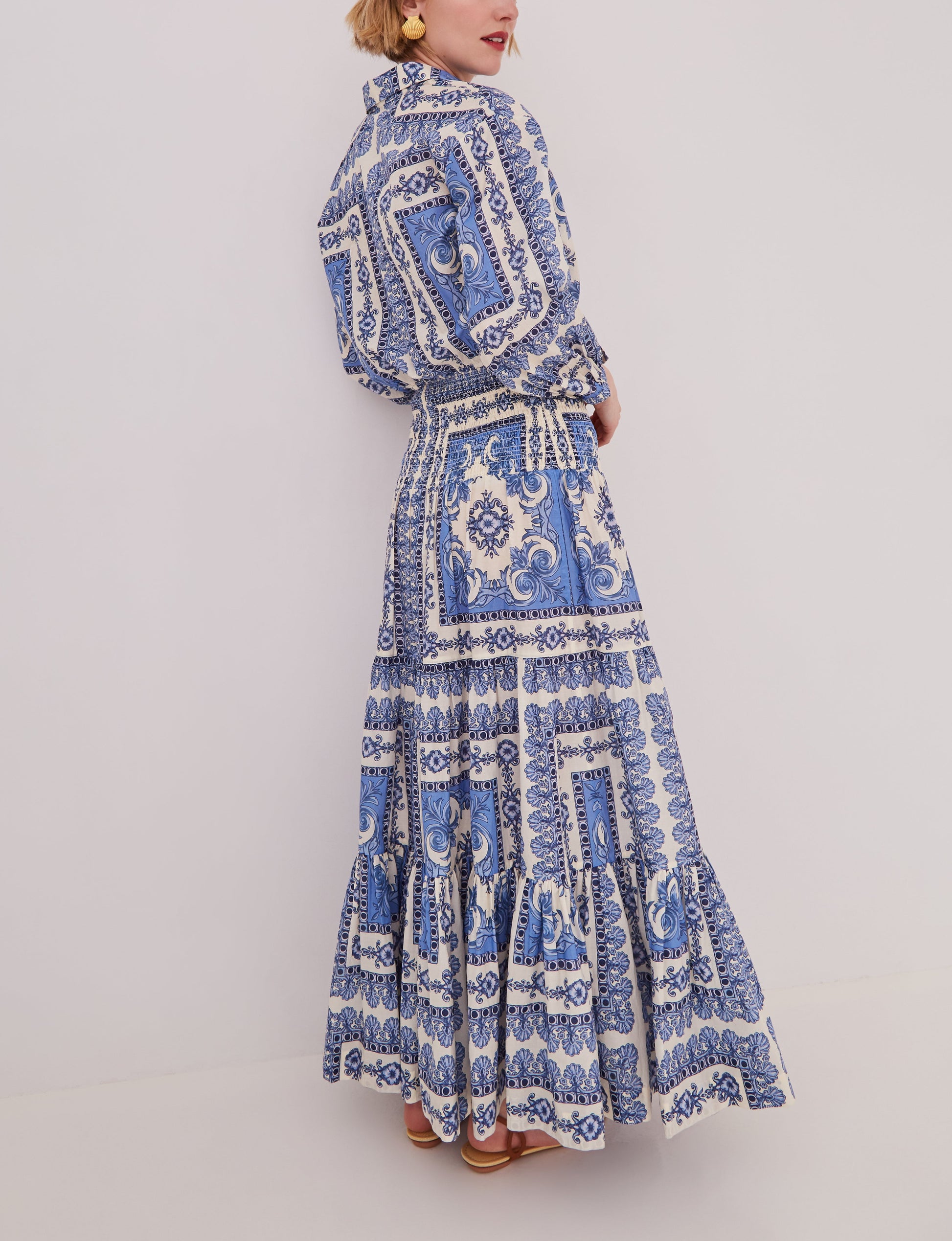 A woman models the Cara Cara Emilia Skirt, a blue and white patterned, voluminous ankle-length skirt, standing against a plain light background and facing slightly away from the camera.