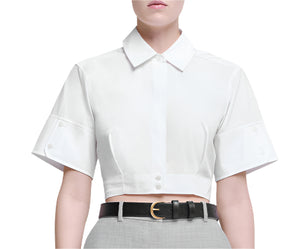 Wearing the Sabrina Top by Simkhai—a white, short-sleeved cropped button-down—paired with grey high-waisted pants and a black belt with a gold buckle for an effortless take on classic tailoring.