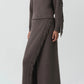 A person wearing a ribbed brown knit sweater and the Simkhai Casey Knit Skirt—a matching wool-cashmere blend fringed maxi skirt with a slit—paired with brown pointed boots.