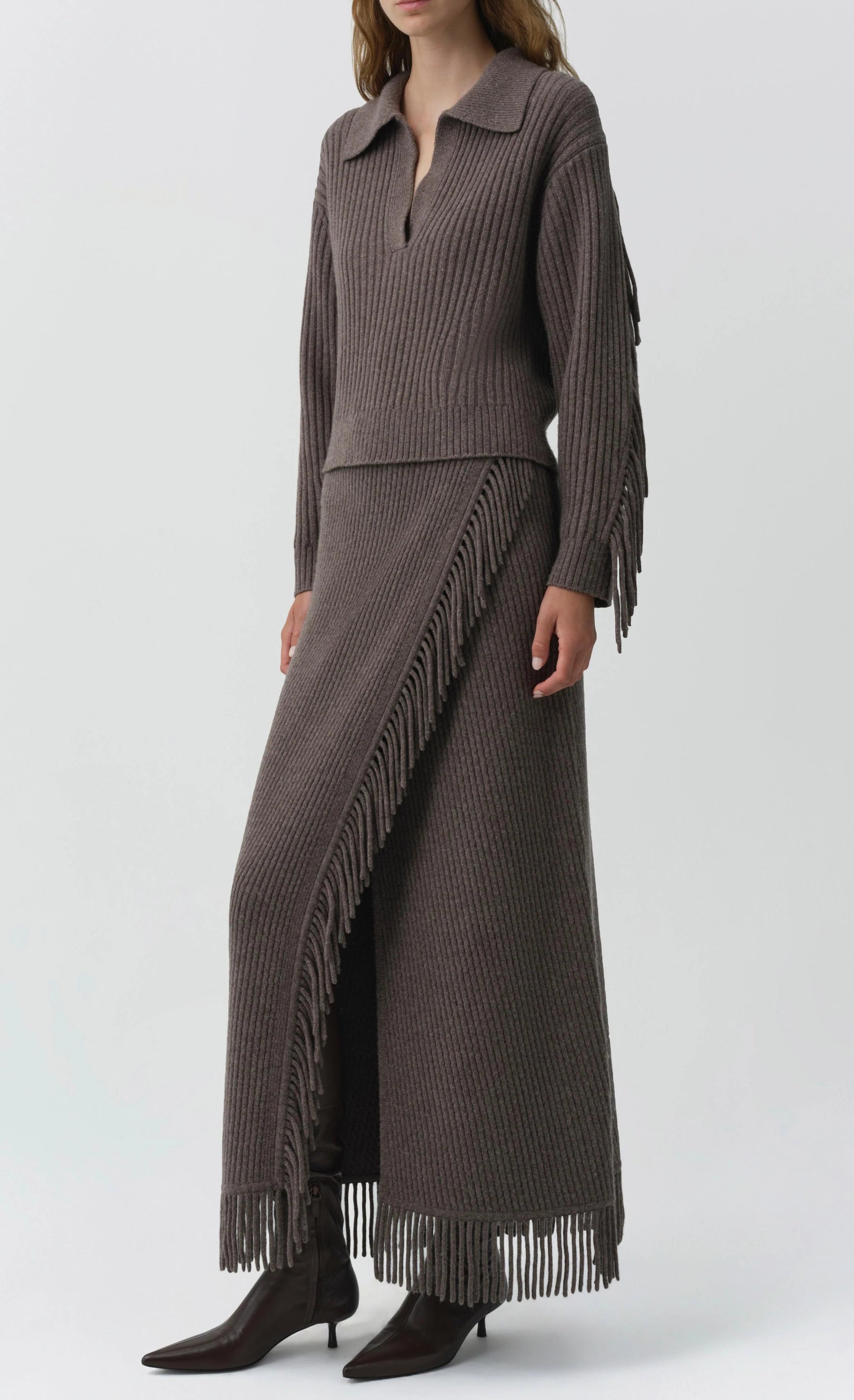 A person wearing a ribbed brown knit sweater and the Simkhai Casey Knit Skirt—a matching wool-cashmere blend fringed maxi skirt with a slit—paired with brown pointed boots.