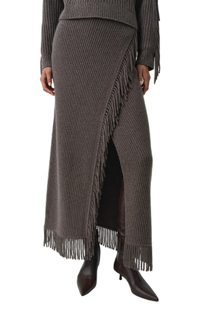 A person is seen wearing the Simkhai Casey Knit Skirt—a ribbed midi skirt featuring fringe details at the hem and a faux-wrap design, crafted from a luxurious wool-cashmere blend—paired with heeled boots; only the upper body is partially visible.