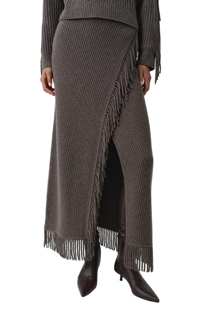 A person is seen wearing the Simkhai Casey Knit Skirt—a ribbed midi skirt featuring fringe details at the hem and a faux-wrap design, crafted from a luxurious wool-cashmere blend—paired with heeled boots; only the upper body is partially visible.
