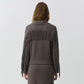 A person faces away in the Simkhai Natalia Sweater—a ribbed, taupe knit with long fringe and Western-inspired detailing across the back and sleeves—paired with a matching skirt, set against a plain light background.