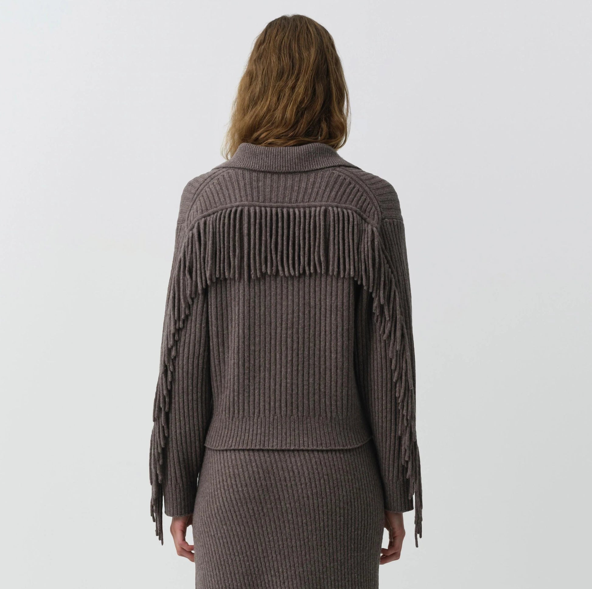 A person faces away in the Simkhai Natalia Sweater—a ribbed, taupe knit with long fringe and Western-inspired detailing across the back and sleeves—paired with a matching skirt, set against a plain light background.