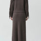 Person wearing the Simkhai Casey Knit Skirt—a long, ribbed, taupe wool-cashmere blend skirt with fringe detailing on the back and hem—shown from behind against a plain background.