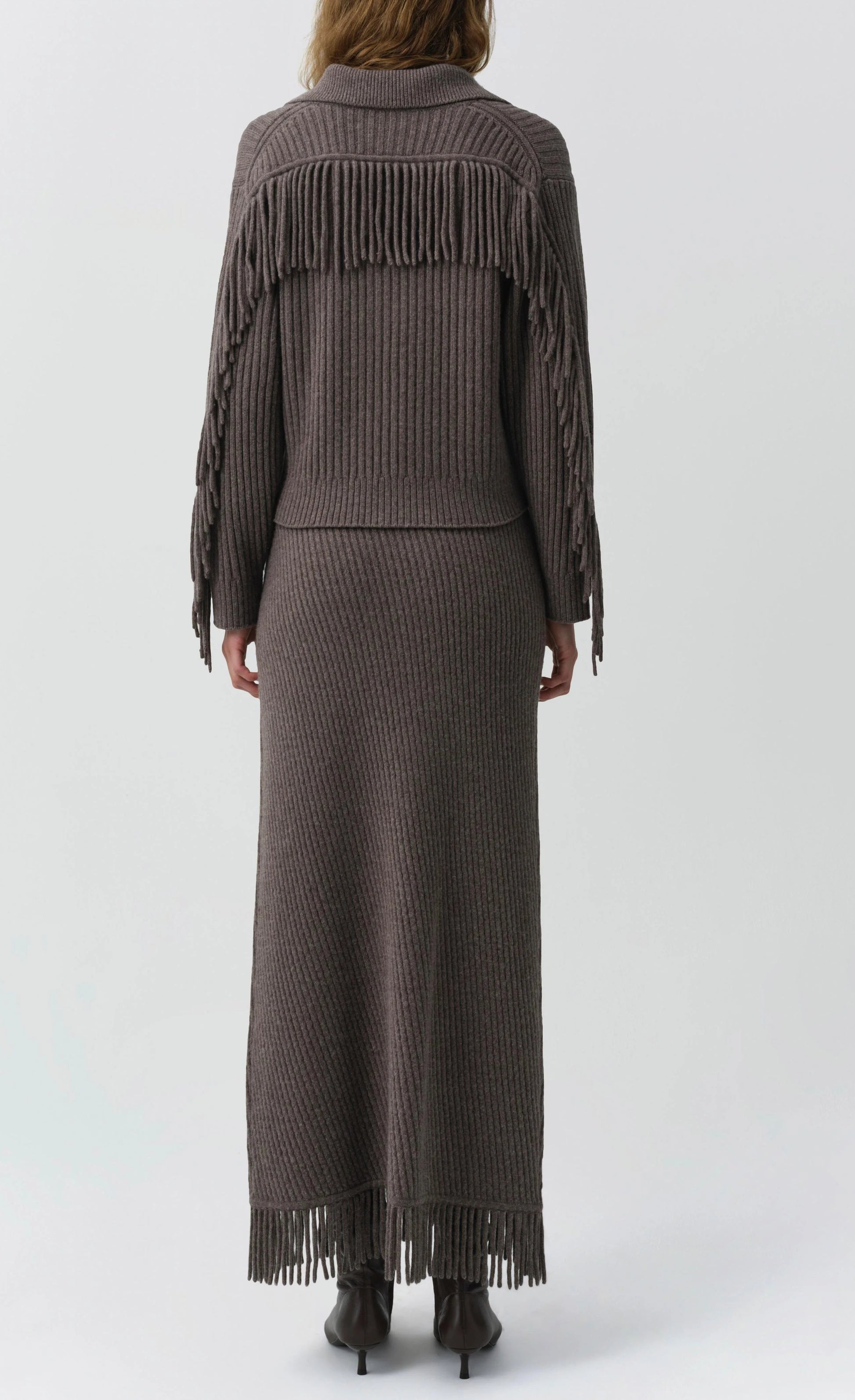 Person wearing the Simkhai Casey Knit Skirt—a long, ribbed, taupe wool-cashmere blend skirt with fringe detailing on the back and hem—shown from behind against a plain background.