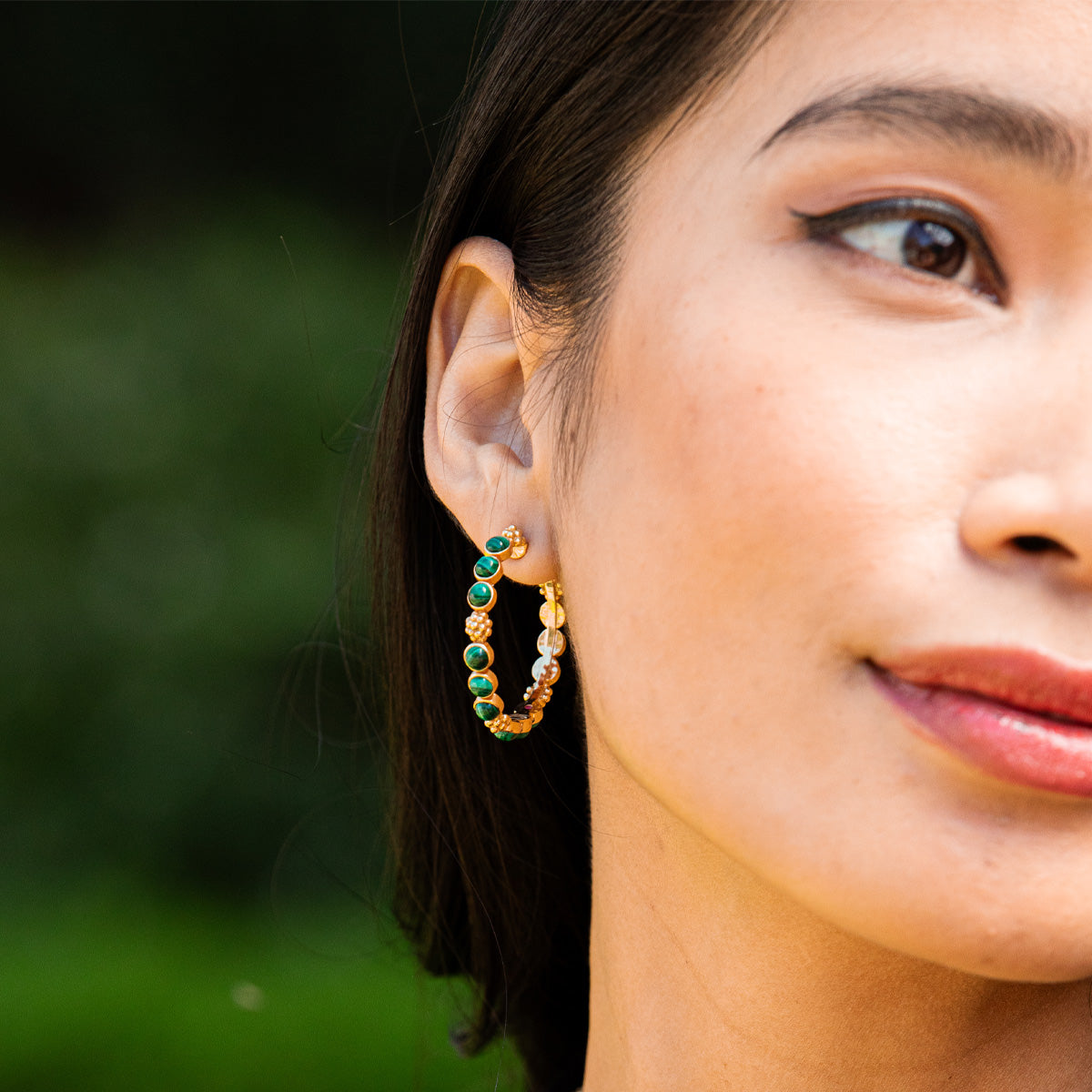 A woman wears Juliska Capucine De Wulf's Berry Gem Medium Hoop Earrings, featuring green and white beads, against a blurred outdoor background.