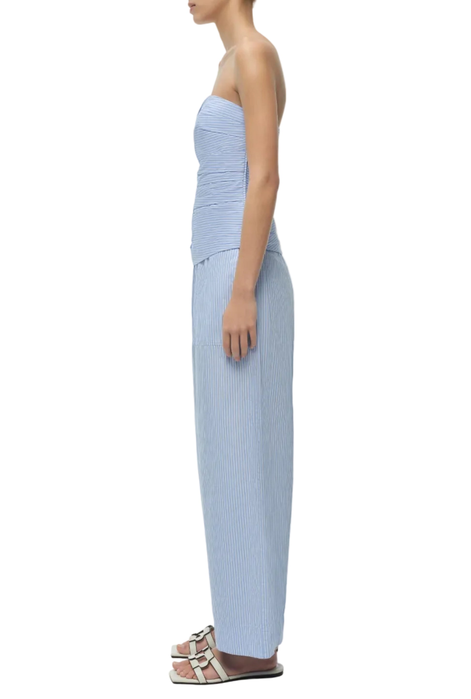 A person stands in profile against a white background, wearing Simkhai’s Arden Pull On Pant—a strapless, light blue pinstriped cotton set with matching wide-leg pants and white sandals.