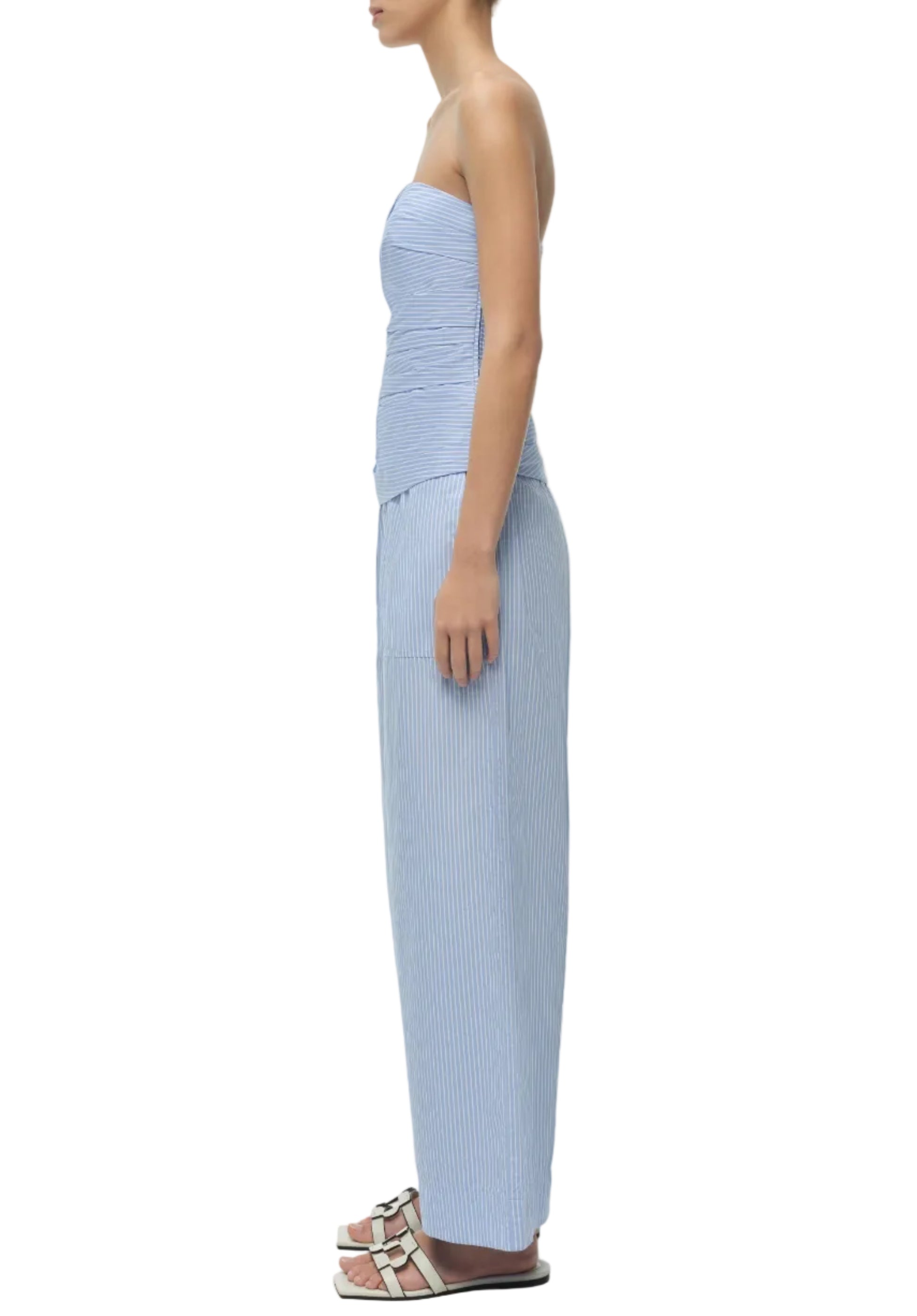 A woman stands in profile wearing the Simkhai Emerson Ruched Bustier Top in light blue, paired with matching high-waisted, wide-leg pants and white slide sandals.