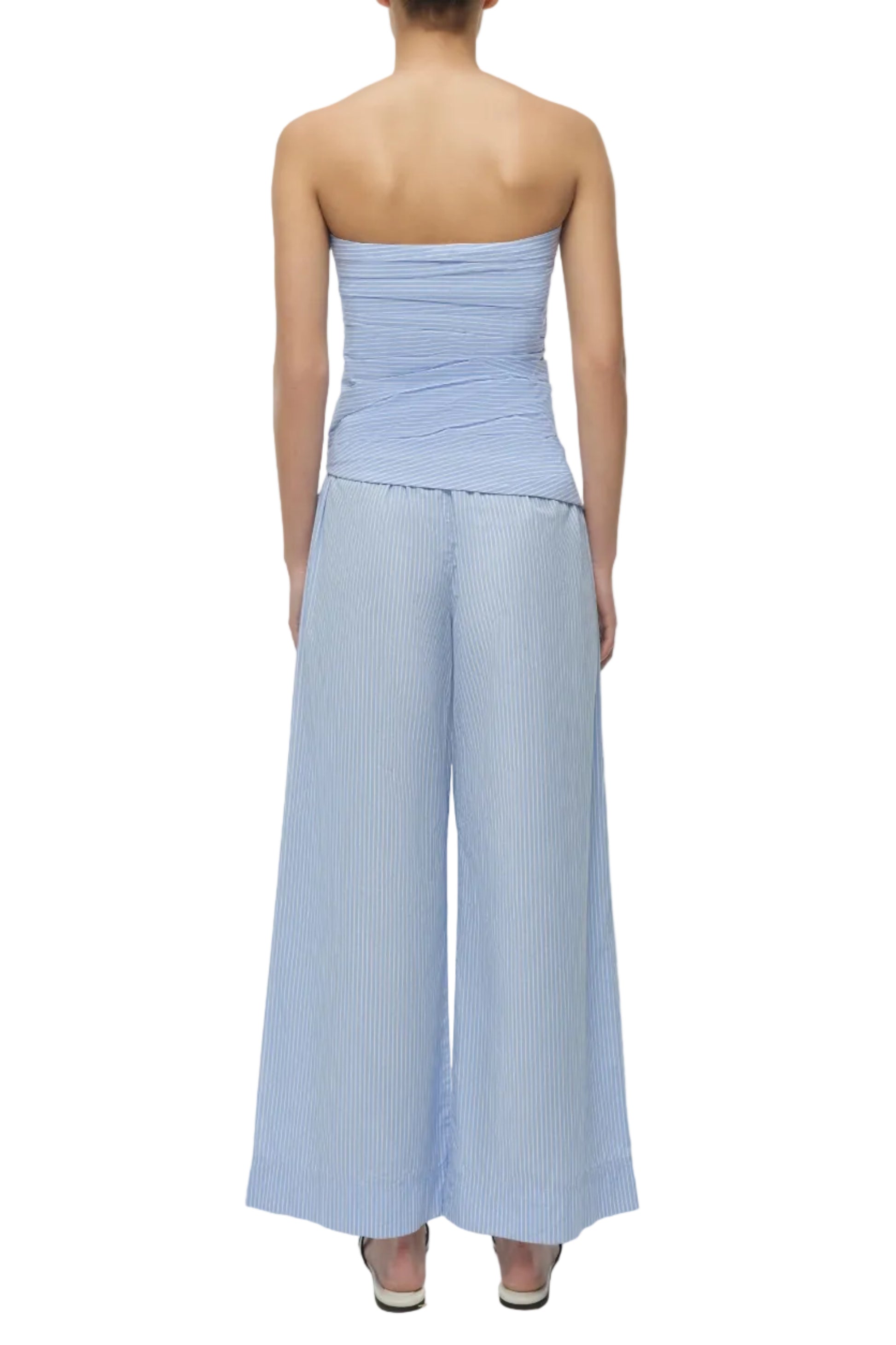 A woman is seen from behind wearing Simkhai’s Arden Pull On Pant, a light blue and white striped, wide-leg style crafted from lightweight cotton, paired with high heels.