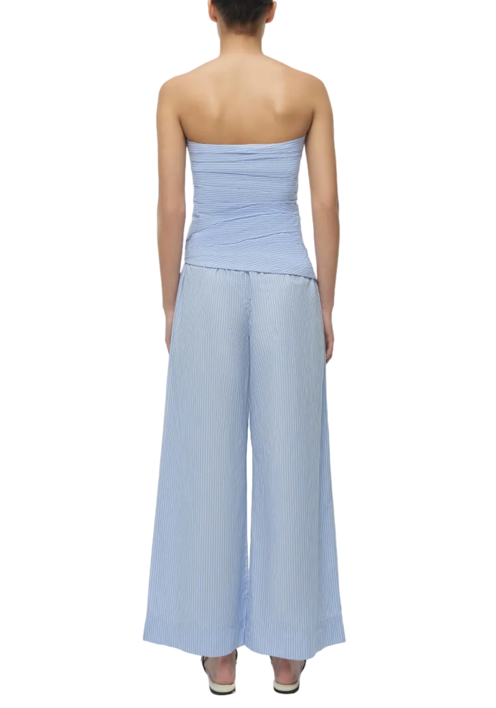 A person faces away in the Simkhai Emerson Ruched Bustier Top, a strapless light blue piece, paired with matching wide-leg striped cotton poplin pants and heels.