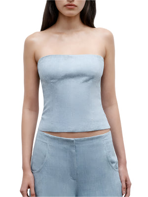 A woman wearing the TWP Jillian Bustier, a light blue strapless top, paired with matching high-waisted hemp denim pants featuring side pockets.