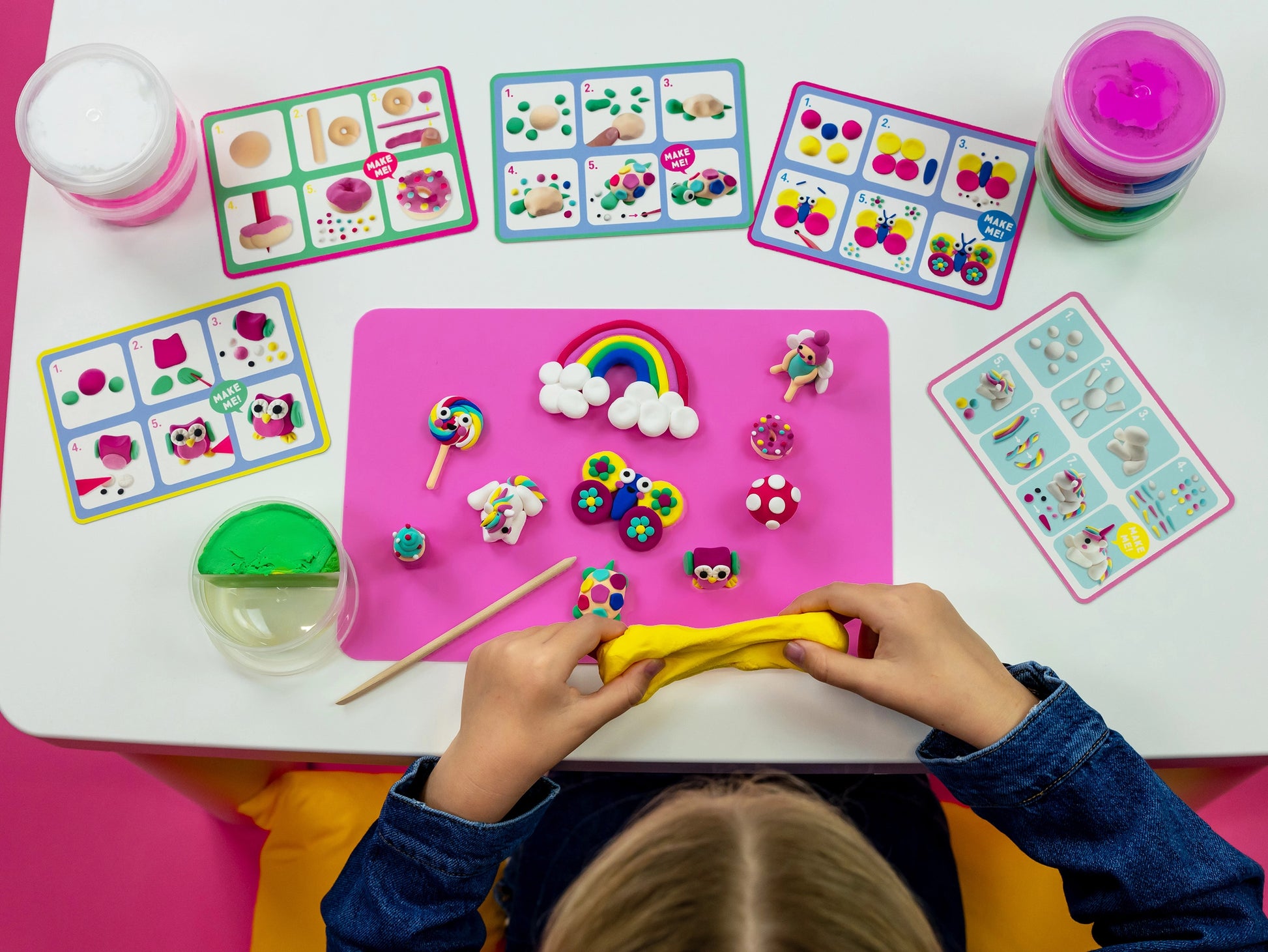 A child molds yellow playdough at a table with cards displaying clay models, finished figures, and containers of colored playdough from the Princess Clay & Play sculpting kit by Floss and Rock.