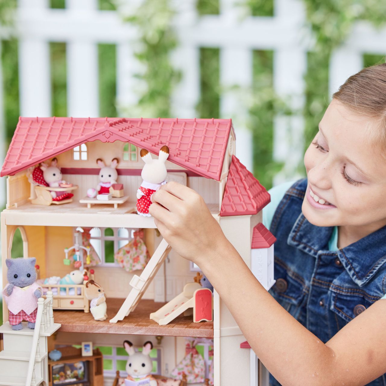 A child smiles while playing with Calico Critters animal figurines from the Chocolate Rabbit family in a large Country Home-Secret Attic Playroom dolls house, set outdoors with a white picket fence in the background.