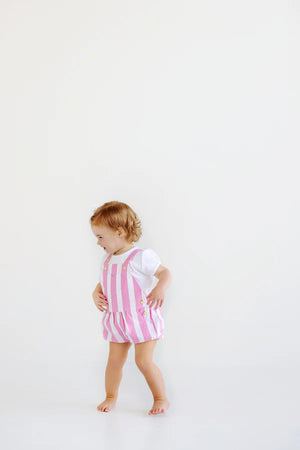 A toddler wearing a white Baby Girls' Penny's Play Onesie from The Beaufort Bonnet Company stands barefoot against a plain white background.