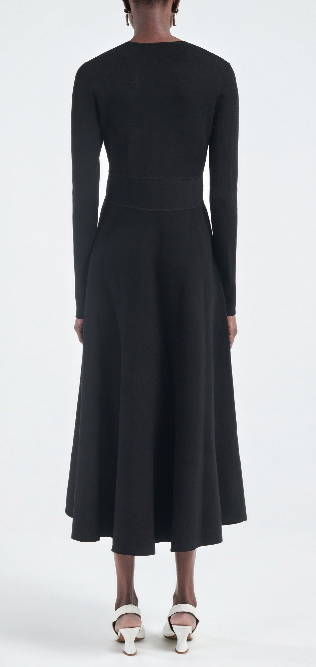 A person faces away, wearing the Maria McManus Button Front Dress—an ankle-length, long-sleeved black dress made from FSC-certified viscose—with white heeled shoes against a plain light backdrop.